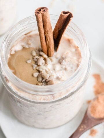 Overnight oats in a jar with cinnamon, seeds, and seed butter next to a spoonful of cinnamon.