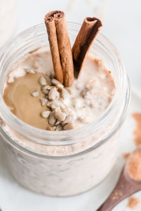 Overnight oats in a jar with cinnamon, seeds, and seed butter next to a spoonful of cinnamon.