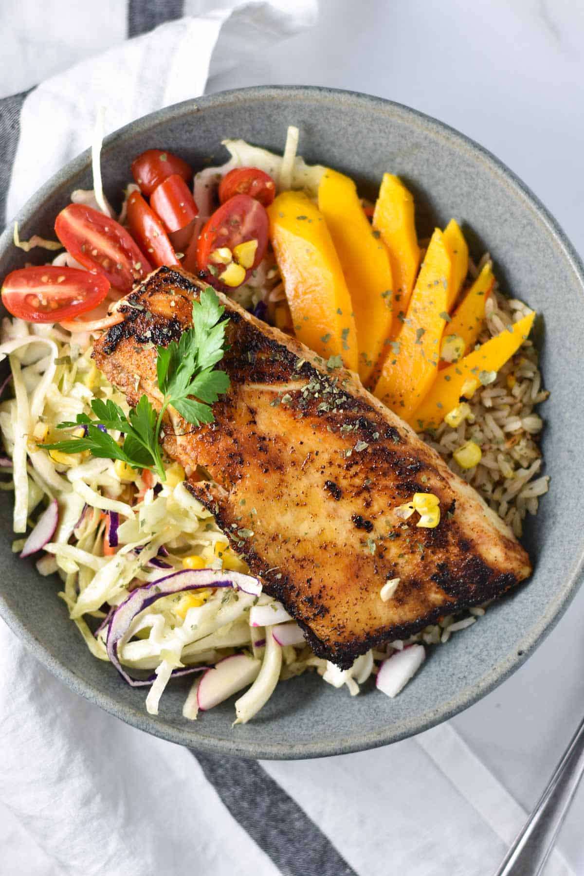 A gray bowl filled with slaw, tomatoes, mango, fish, and blackened fish.