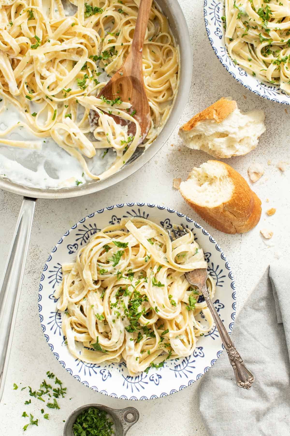 A bowl of fettuccine alfredo next to a baguette and a stainless steel pan of more pasta.