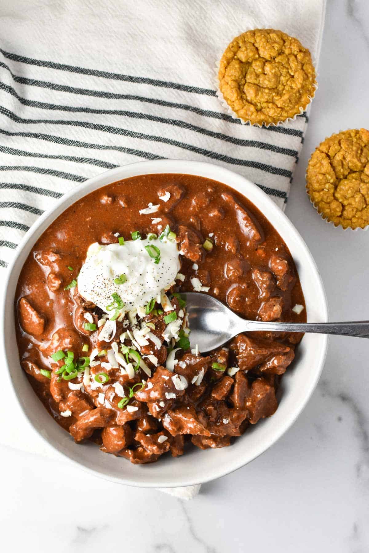 A bowl of Texas chili topped with green onion and next to cornbread muffins.