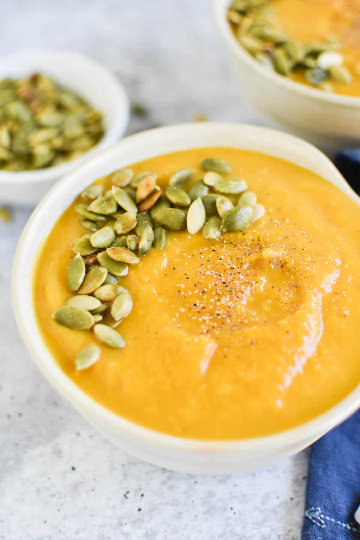 A large bowl of orange autumn squash soup topped with pepitas on a blue napkin.
