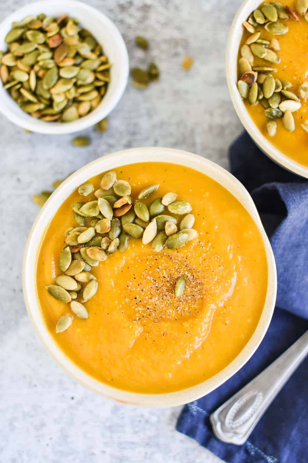Two butternut squash soups topped with pumpkin seeds on a grey background. 