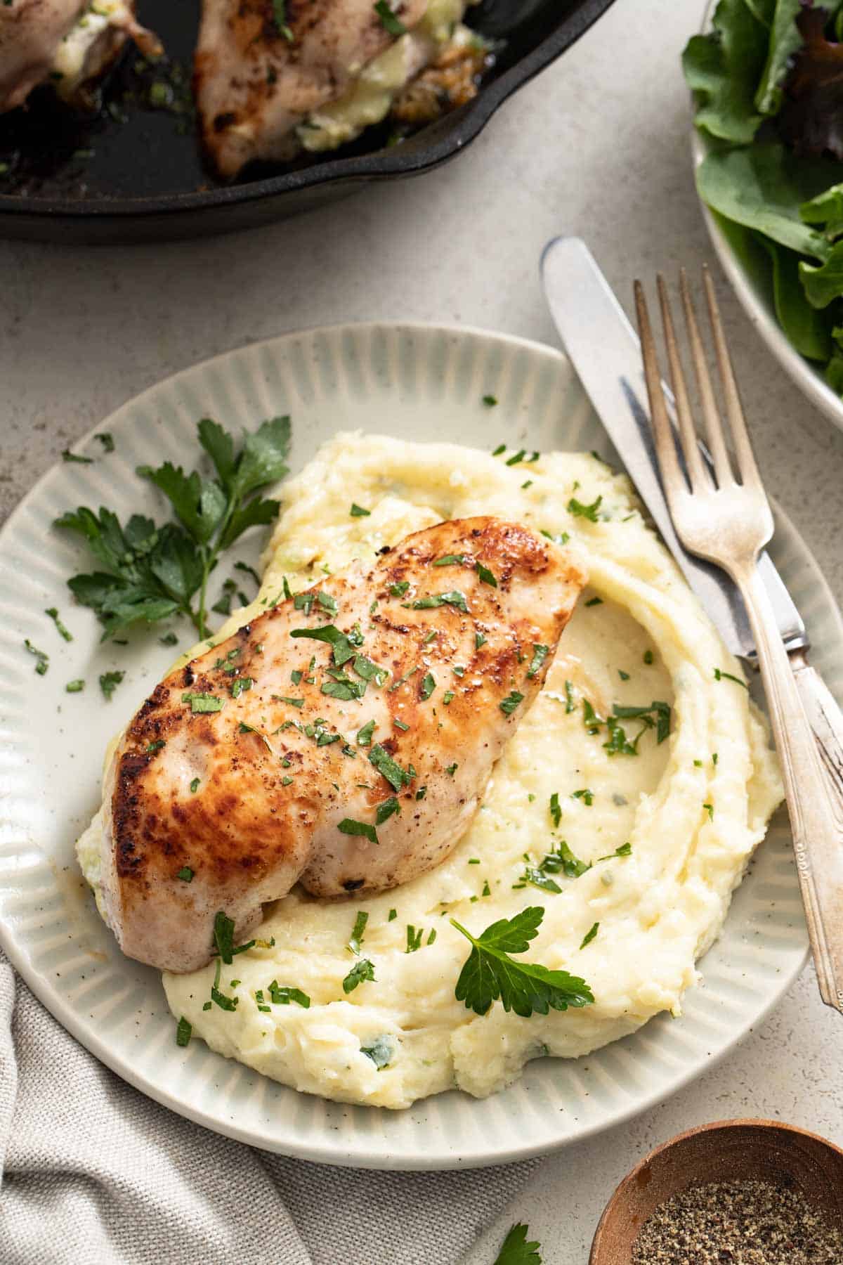 A stuffed chicken breast on top mashed potatoes next to a cast iron pan and parsley. 