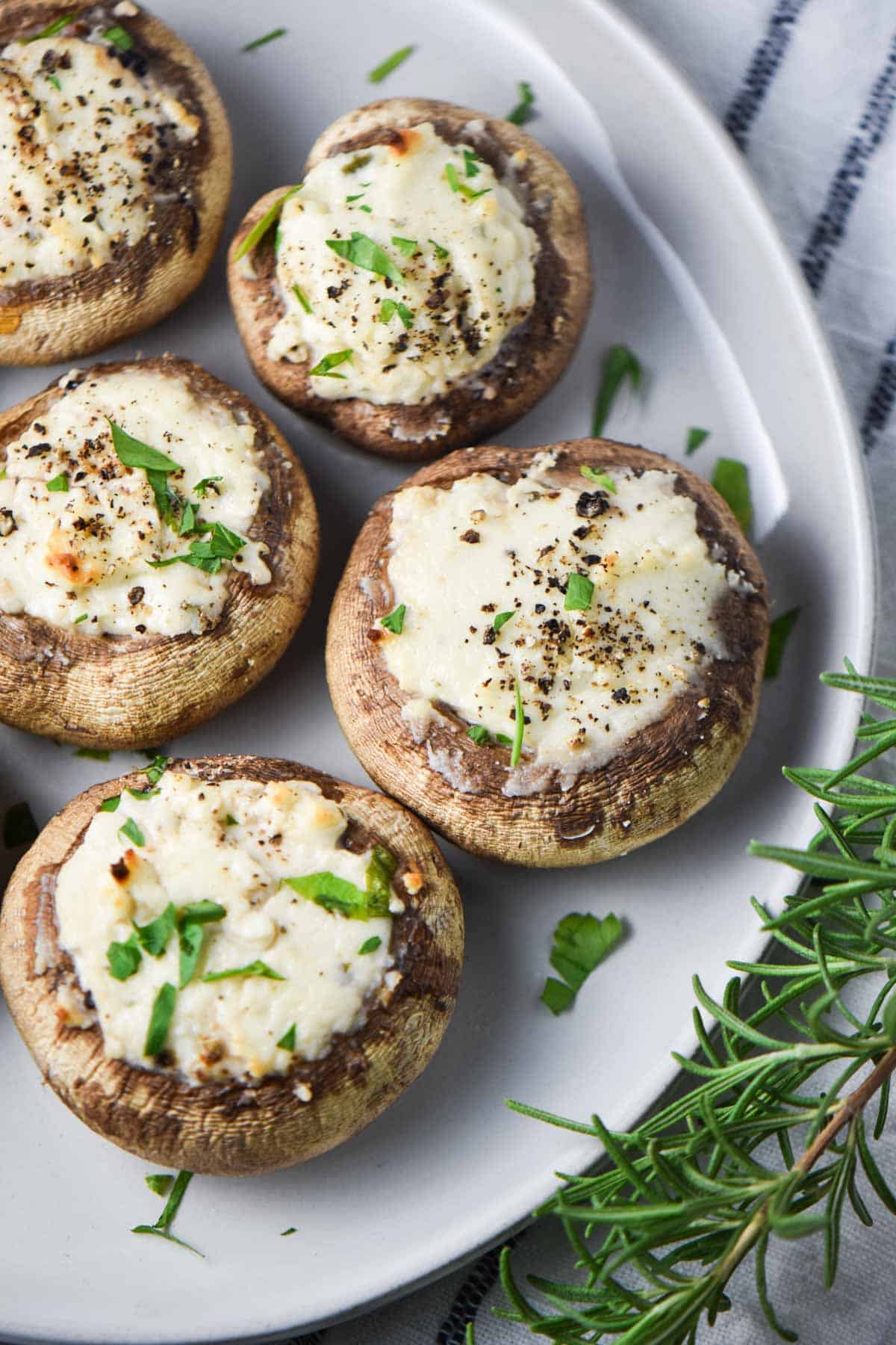 Baked stuffed mushrooms topped with pepper and parsley next to rosemary leaves on a plate.