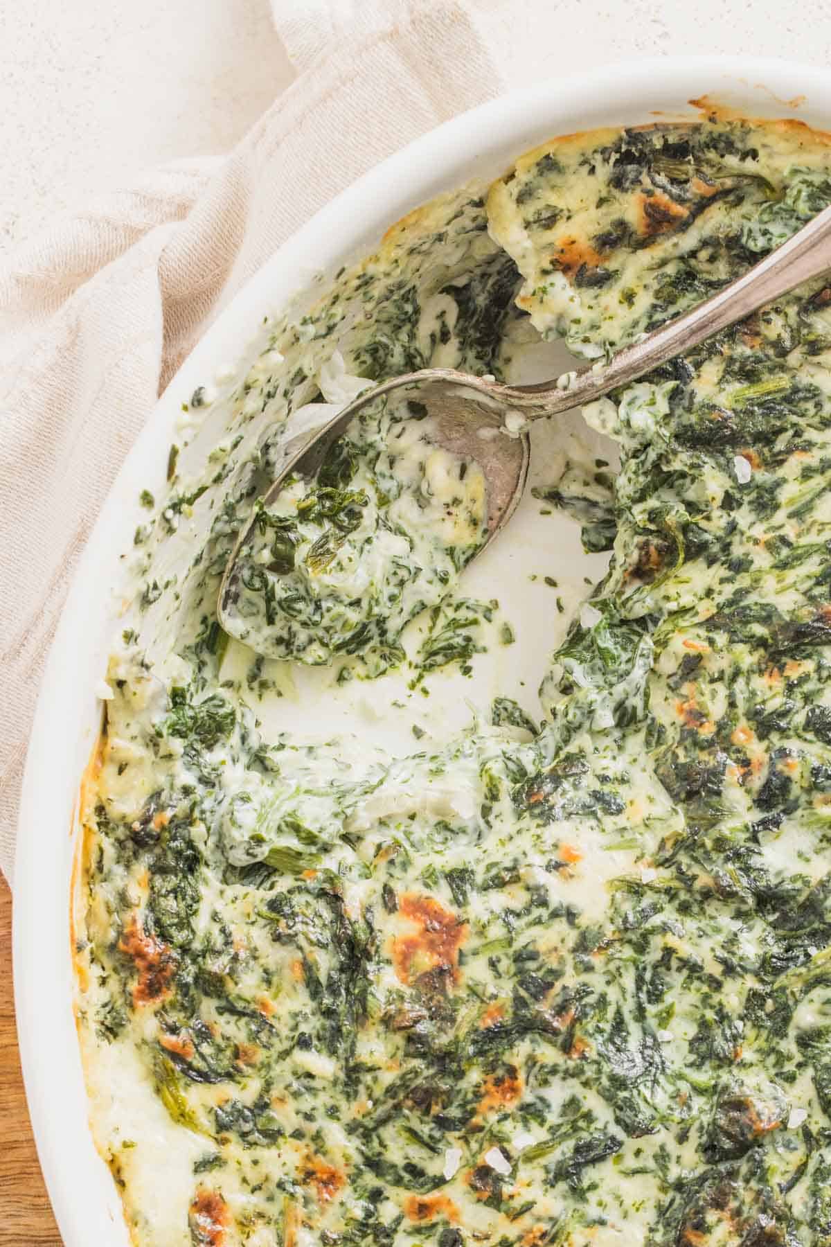 A casserole dish with a silver spoon filled with cheesy creamed spinach.