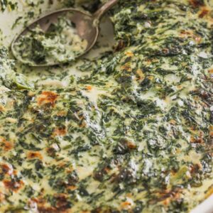 A casserole dish with a silver spoon filled with cheesy creamed spinach.