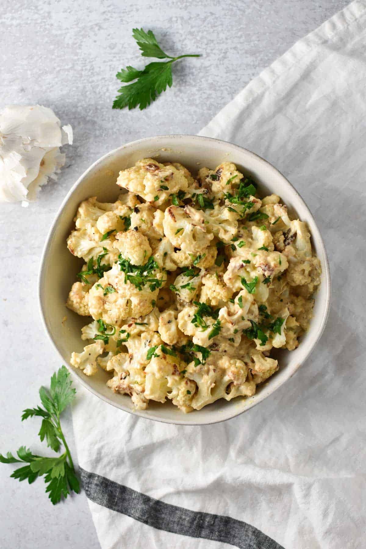 Creamy roasted cauliflower coated in a cheese sauce topped with parsley next to a garlic clove and white napkin.