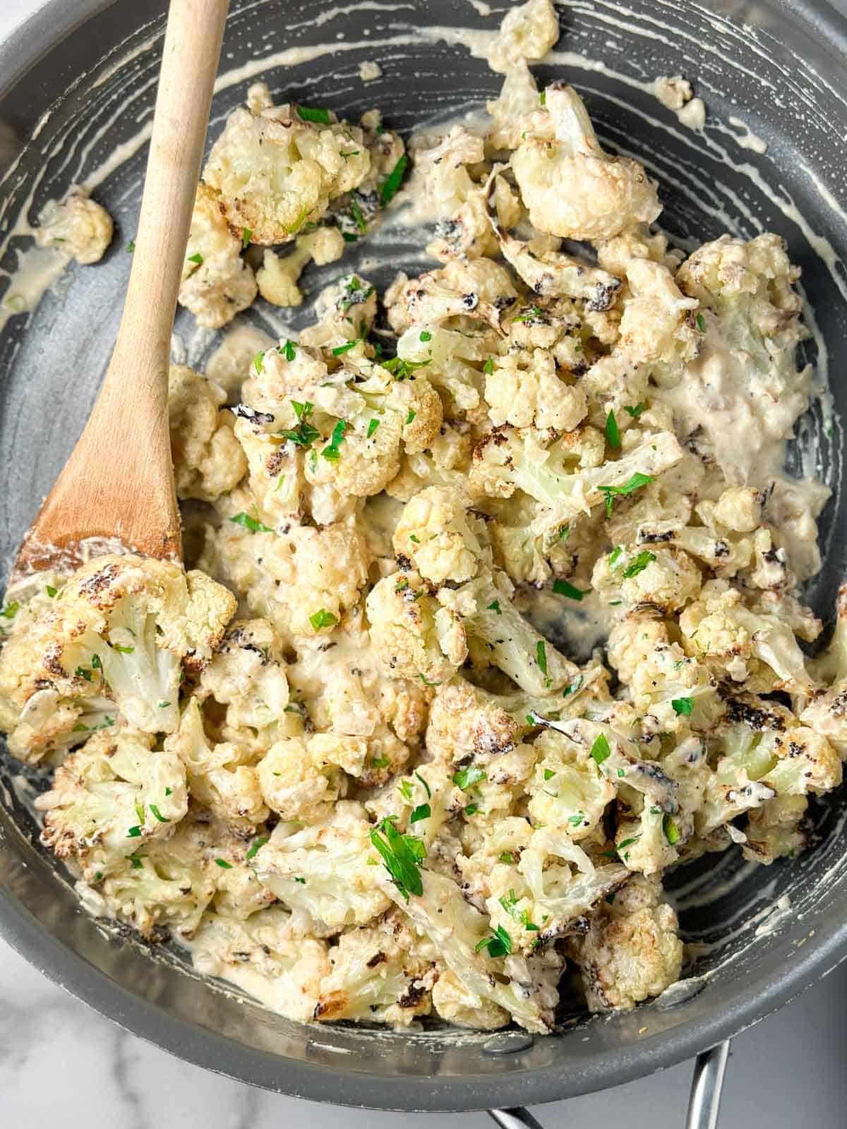 A wood spoon stirring roasted cauliflower in a pan with a cheese sauce.