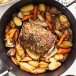 A pork loin roast topped with thyme and surrounded by roasted vegetables in a dutch oven.