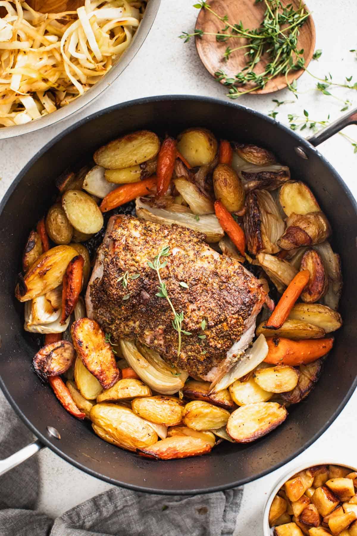 Pork roast in a dutch oven with potatoes and carrots next to a bowl of cooked cabbage and apples. Food is sprinkled with thyme for garnish. 