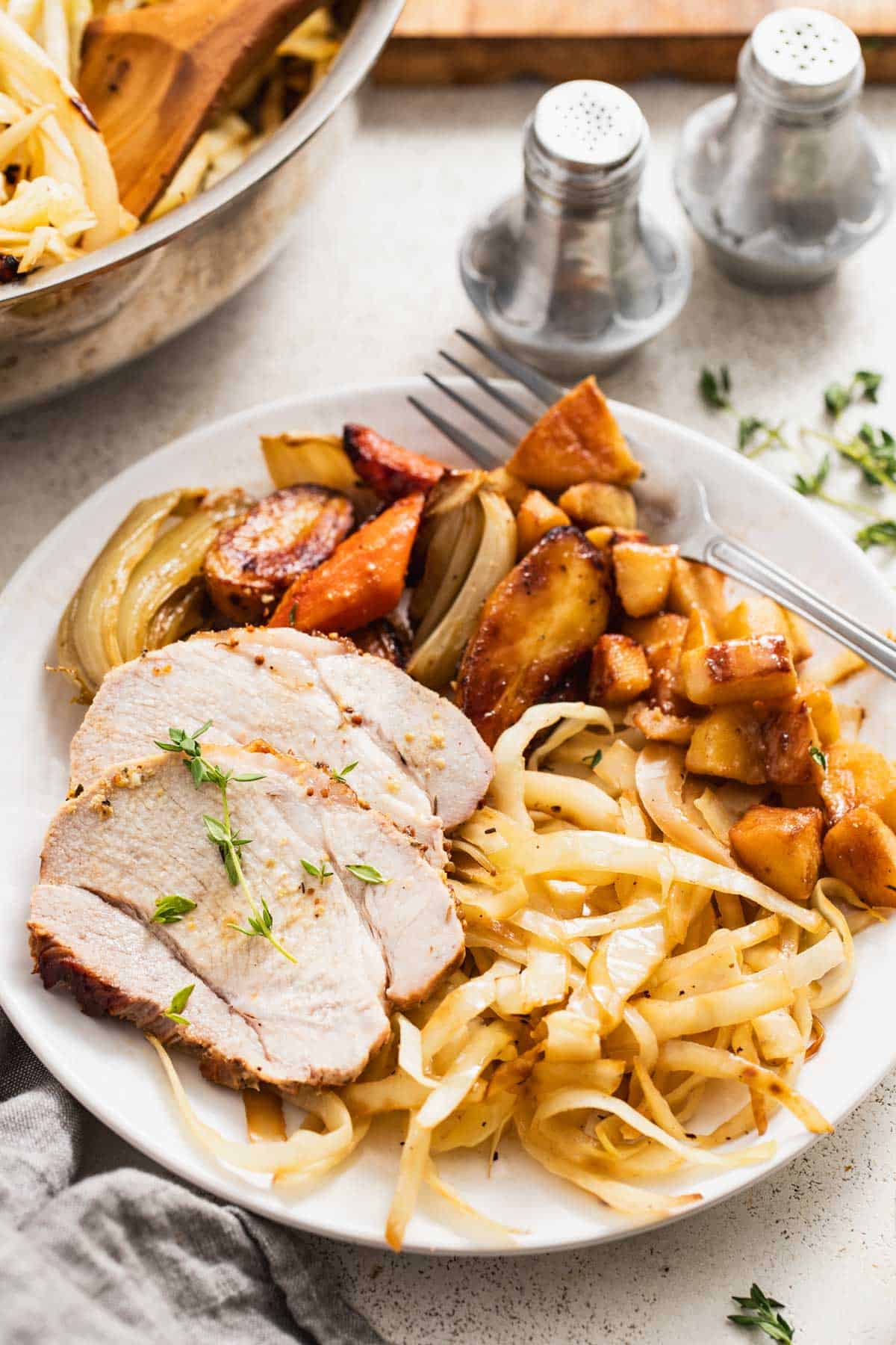 Sliced roasted pork next to cabbage, apples, and roasted root vegetables on a white table.