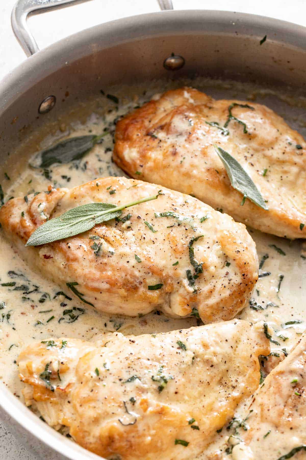 Seared chicken breasts topped in a creamy sauce and sage leaves in a stainless steel pan. 