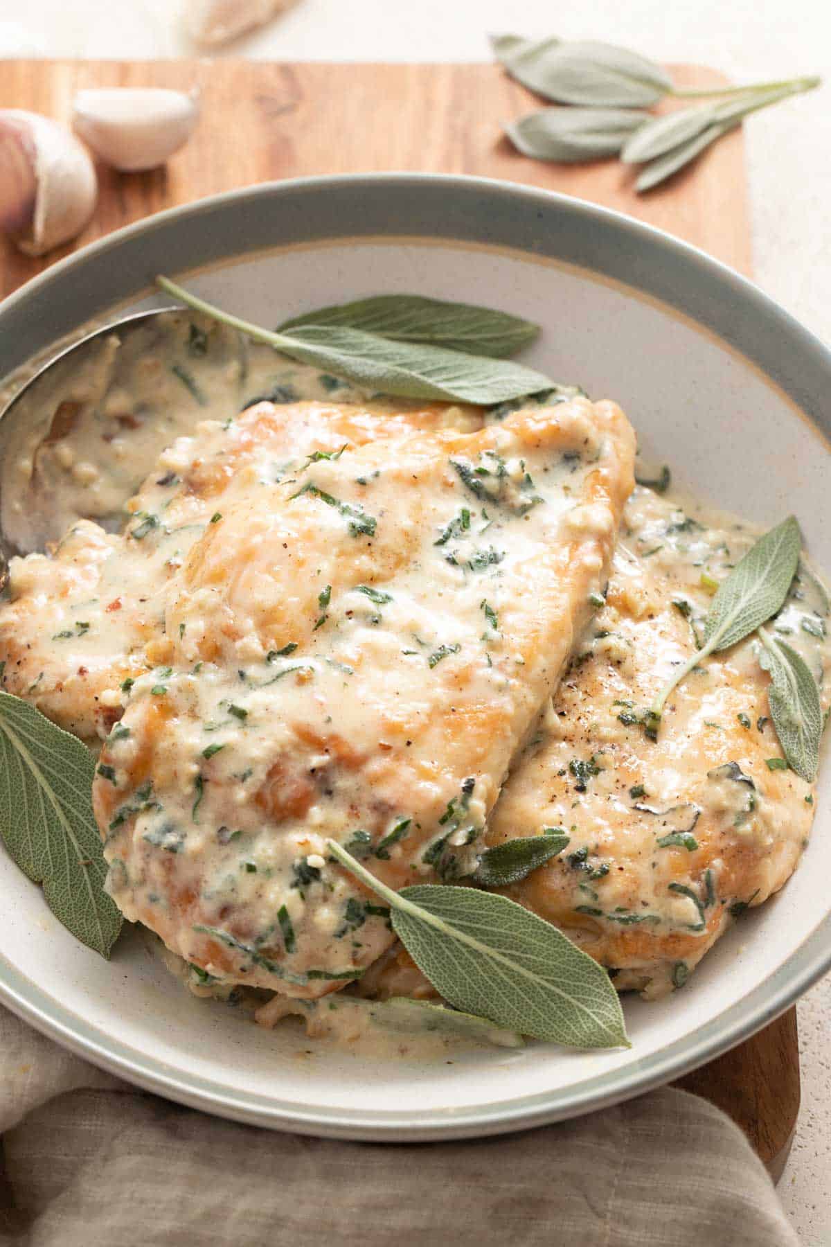 Chicken breasts in a sage cream sauce topped with sage leaves in a gray bowl with garlic cloves next to it.