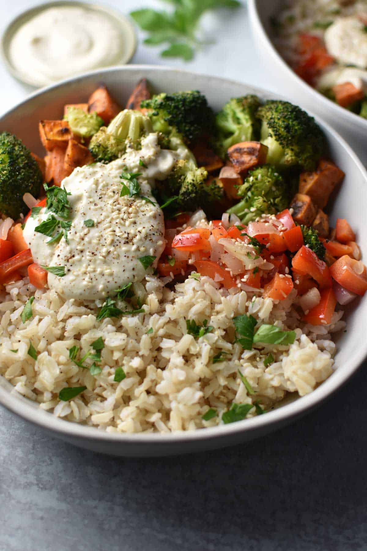 A bowl with sweet potatoes, broccoli, red pepper, rice, and a tahini sauce on top with sesame seeds.