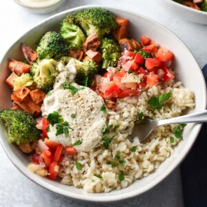 Broccoli, sweet potatoes, and rice all topped with chopped veggies and a sauce with a fork placed on the side of the bowl.