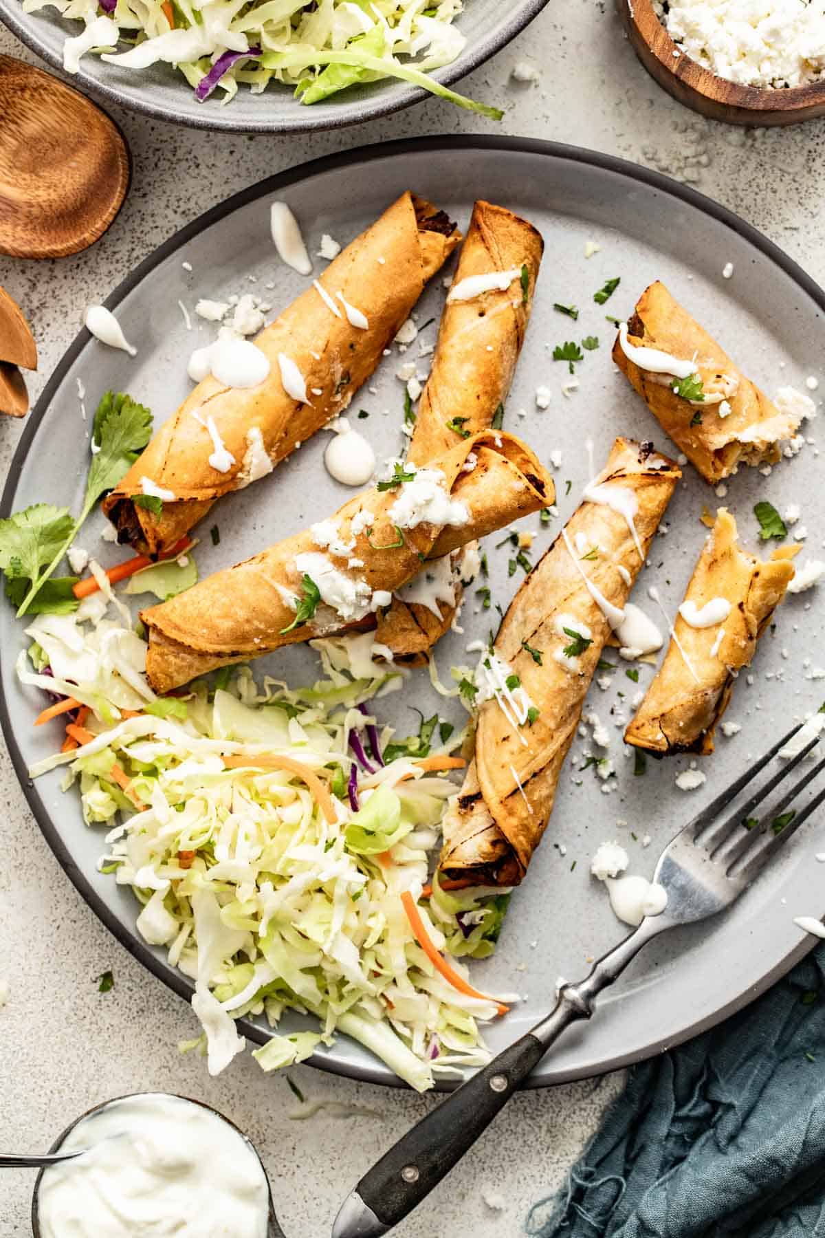 Chicken taquitos stacked next to shredded slaw, a whipped cottage cheese, and cilantro.