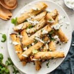 Crispy chicken taquitos on a white plate with sour cream dipping sauce, cilantro, and crumbled cheese.