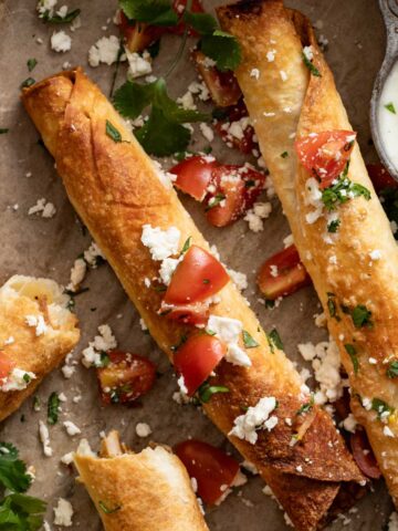 Baked chicken flautas with crumbled cheese, tomatoes, and cilantro on a baking sheet.