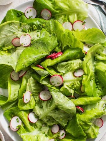 A green butter lettuce side salad topped with sliced radishes, chopped chives, and pepper next to serving utensils and a napkin.
