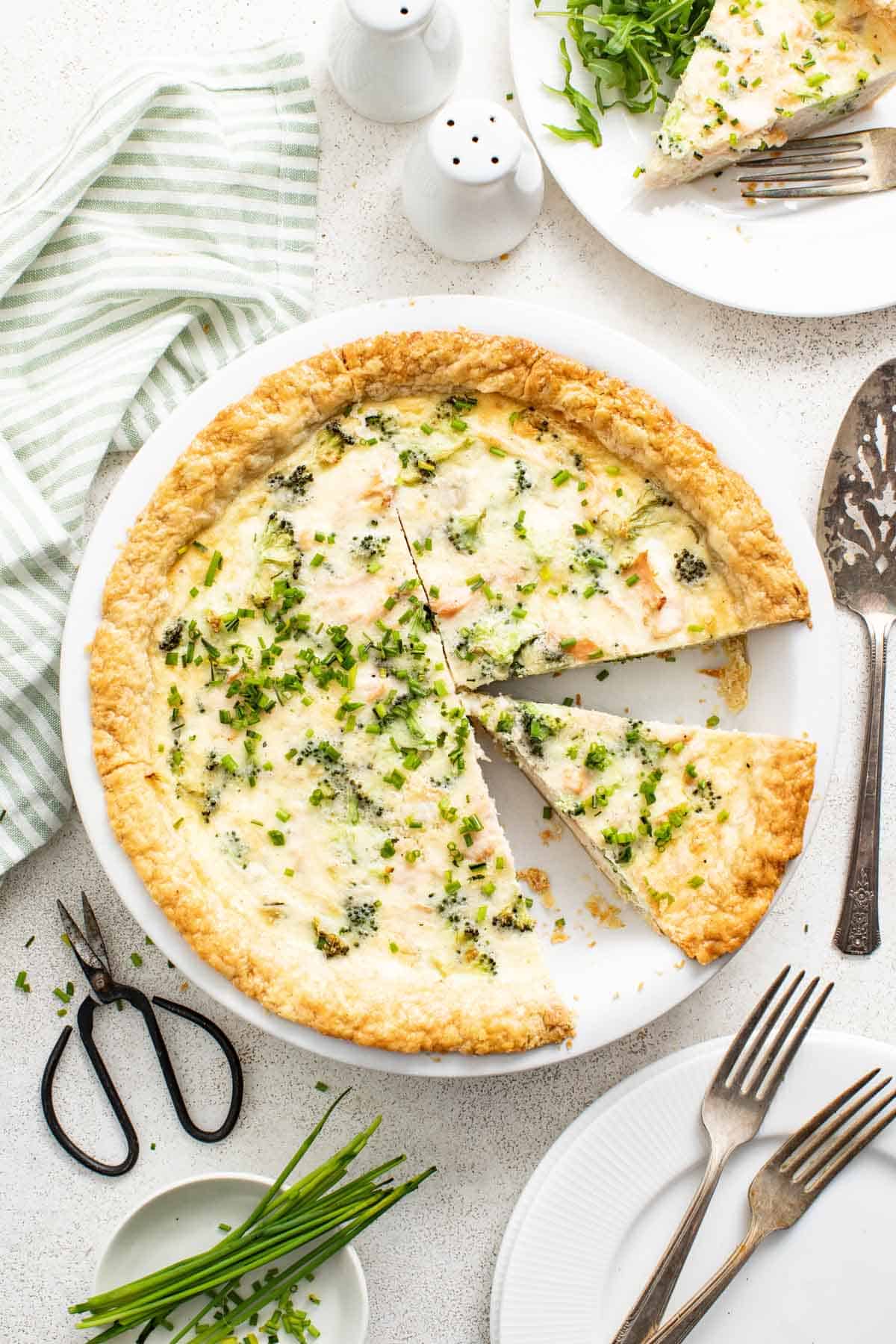 A salmon quiche with broccoli and chives in a white pie plate.