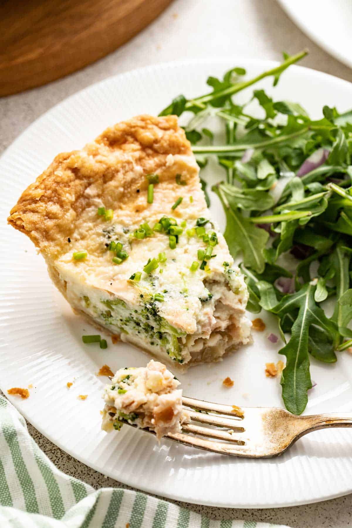 A fork with a bite of quiche with broccoli and salmon on the end on a white plate. 