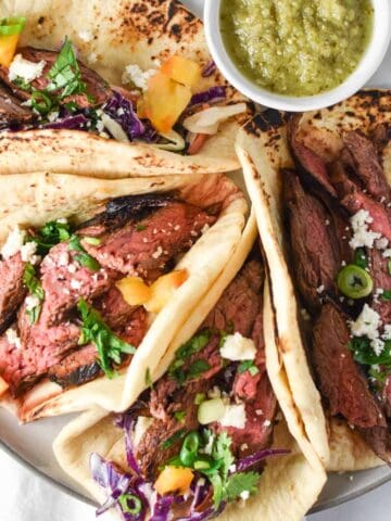 Four flank steak tacos next to a white bowl with salsa verde.