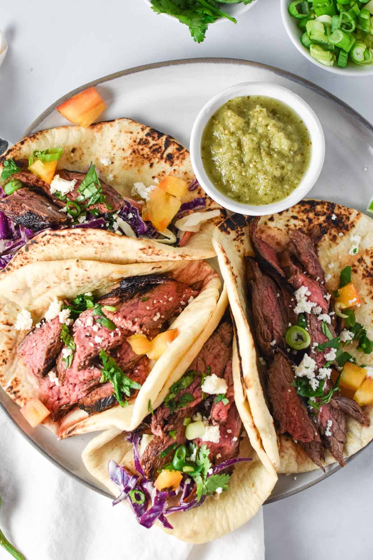 Four steak tacos on a plate with queso fresco and salsa verde.