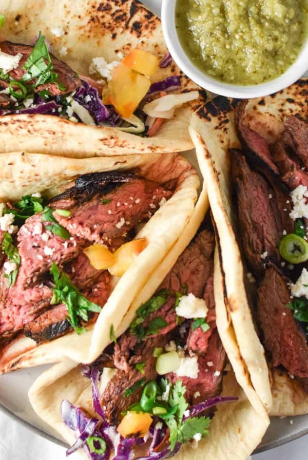 Four flank steak tacos next to a white bowl with salsa verde.