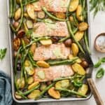 A sheet pan with baked salmon, an herb sauce, green beans and potatoes next to a spatula and salt and pepper.