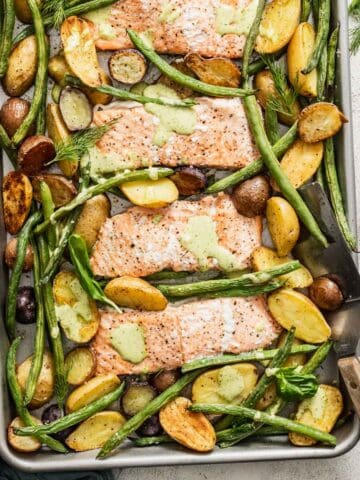 A sheet pan with baked salmon, an herb sauce, green beans and potatoes next to a spatula and salt and pepper.