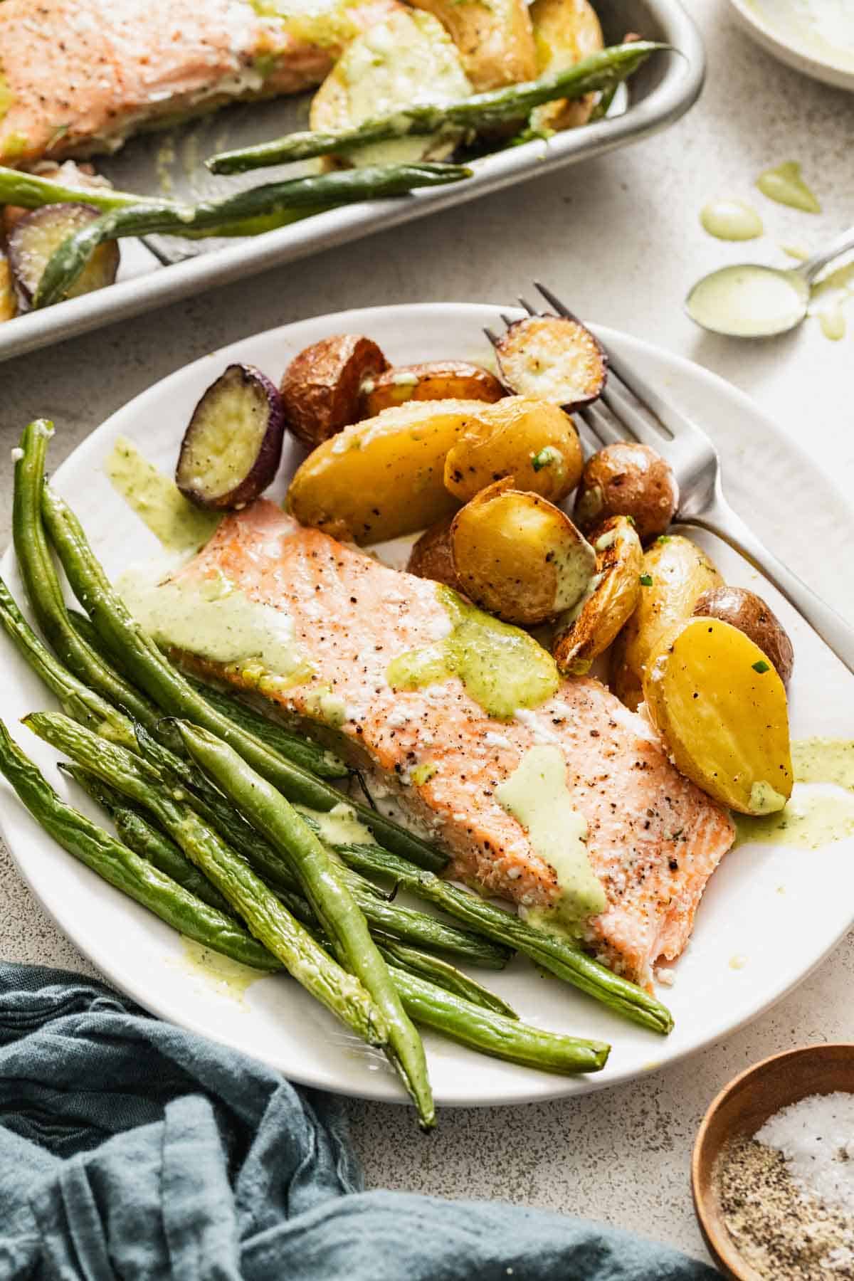 A white plate with salmon, fingerling potatoes, and green beans topped with a green goddess sauce.