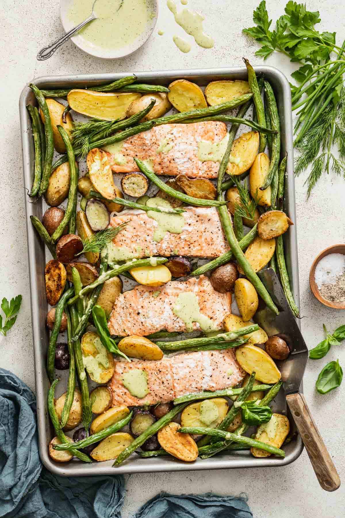 A sheet pan with baked salmon, a sauce, green beans and potatoes next to fresh parsley, green goddess dressing, and a napkin.