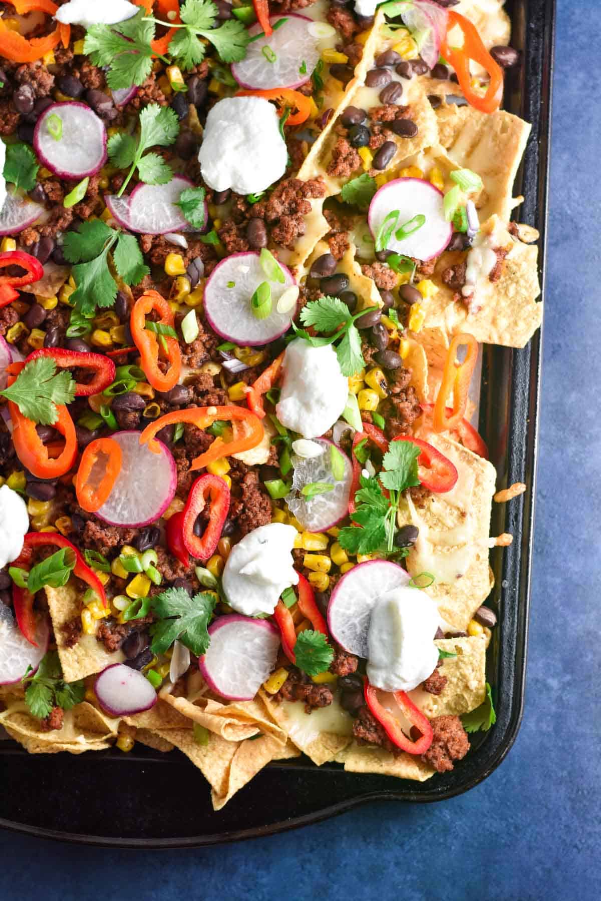 A large sheet pan with tortilla chips, cheese, ground beef, and veggies topped with sour cream.
