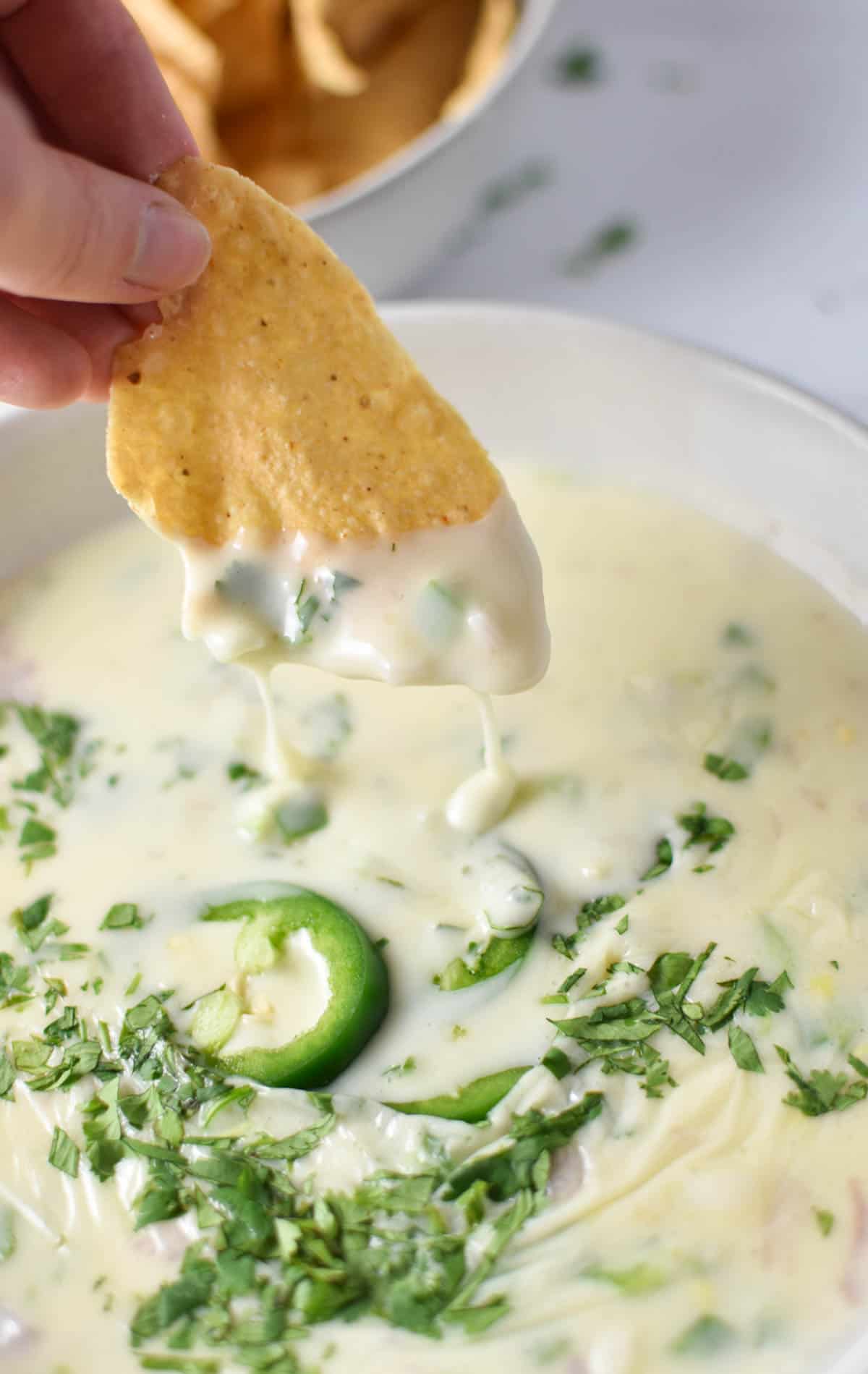 A hand dipping a tortilla chip into a bowl of creamy queso.