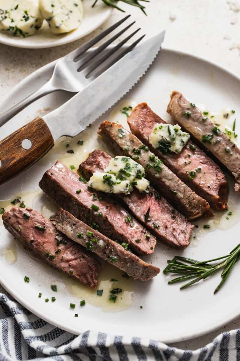 A plate with a fork and knife next to a cooked strip steak that's topped with chives.