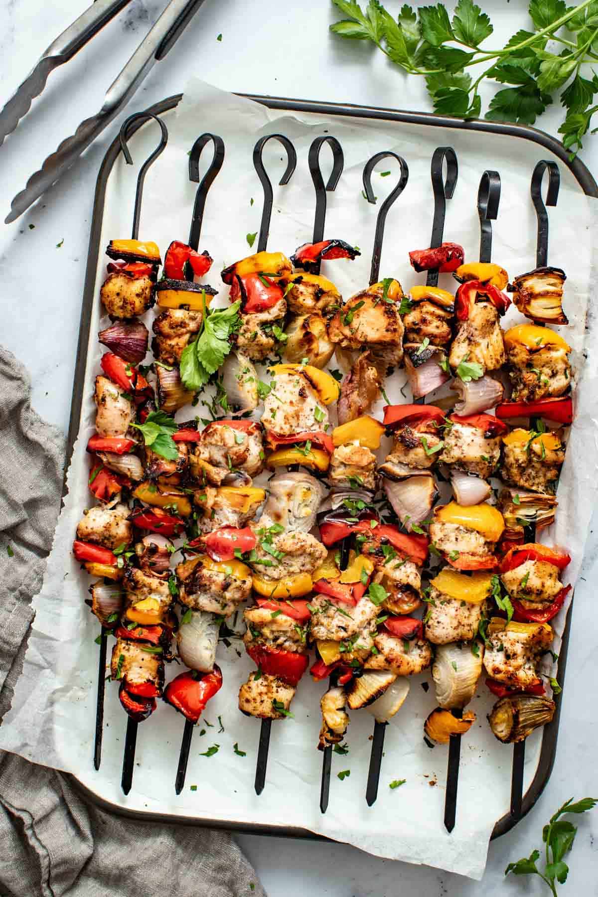 Chicken kabobs on skewers lined up on a baking sheet next to fresh parsley and a grey towel.