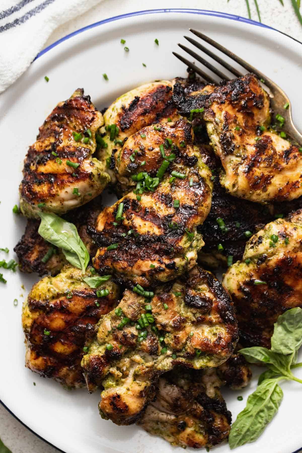 Grilled chicken thighs on a white plate with basil leaves and chives on top.