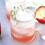 A pink peach mocktail with ice and fresh thyme in a ribbed glass.