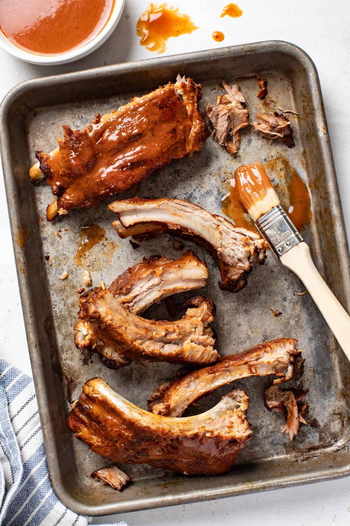 Dry rubbed baby back ribs on a baking sheet, next to a small bowl of homemade bbq sauce and a brush, coating the ribs in the sauce.