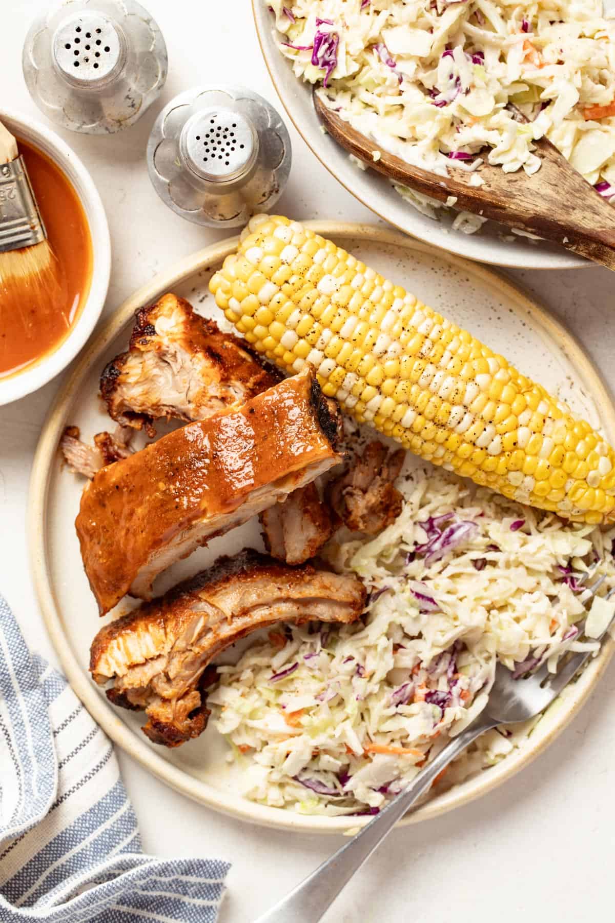 A plate of baby back ribs with dry rub and homemade bbq sauce, next to apple coleslaw and corn on the cob.