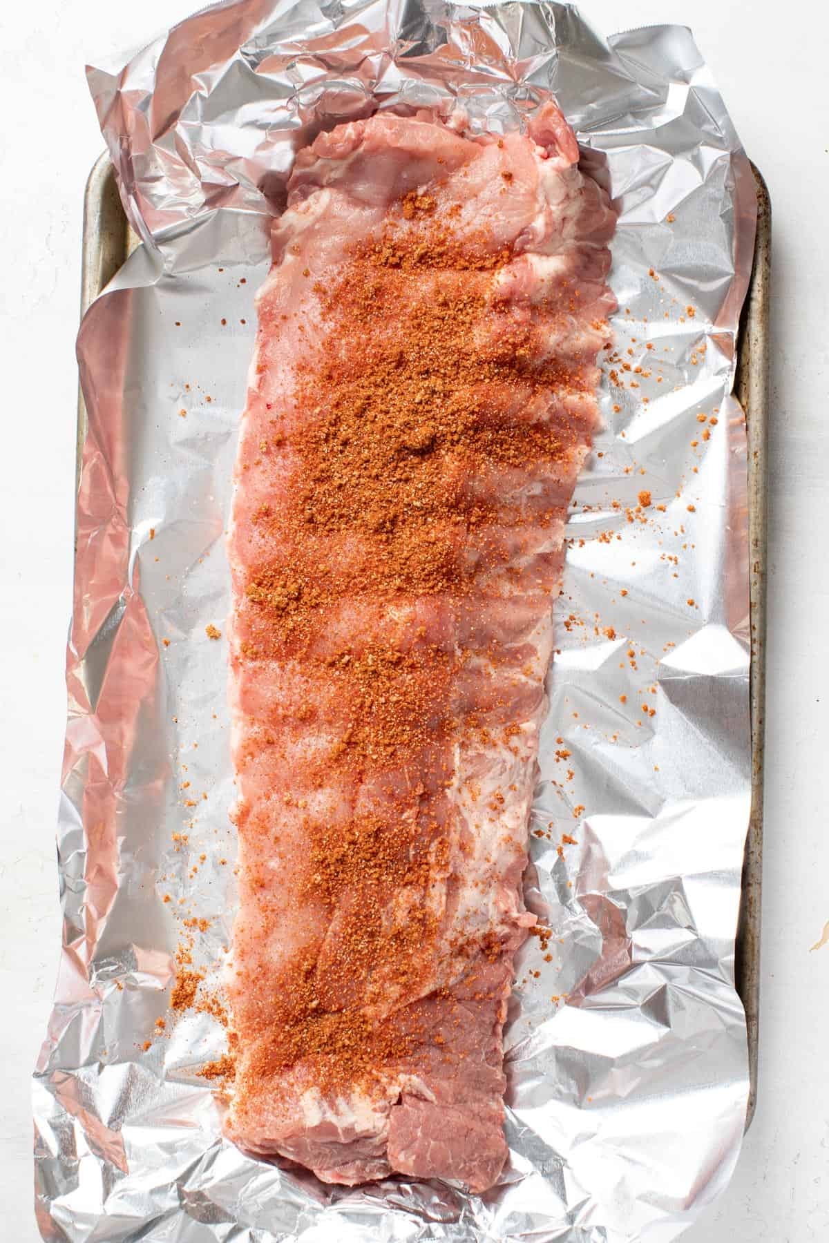 A rack of baby back ribs with a dry rub on top of a sheet of tin foil and baking sheet.