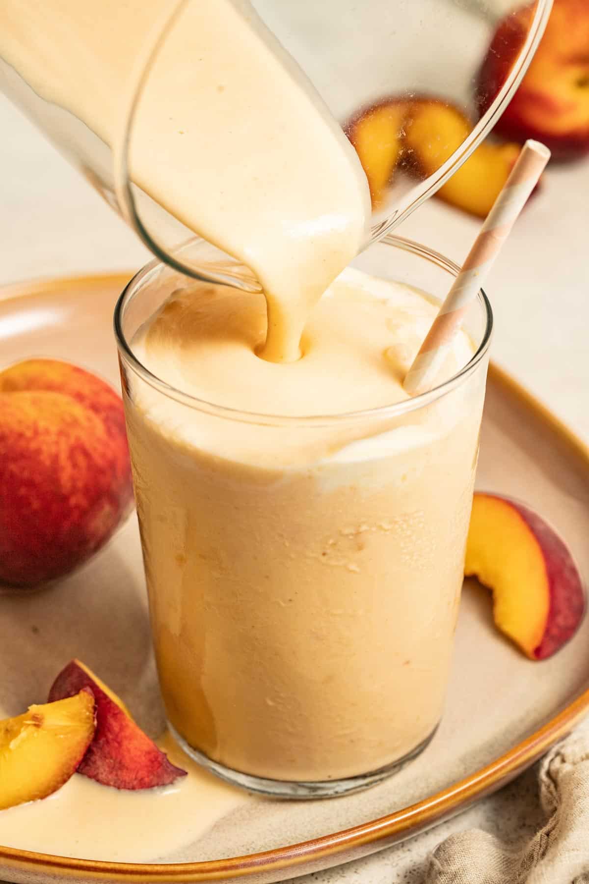 A blender pouring a milkshake with peaches into a glass with a straw.