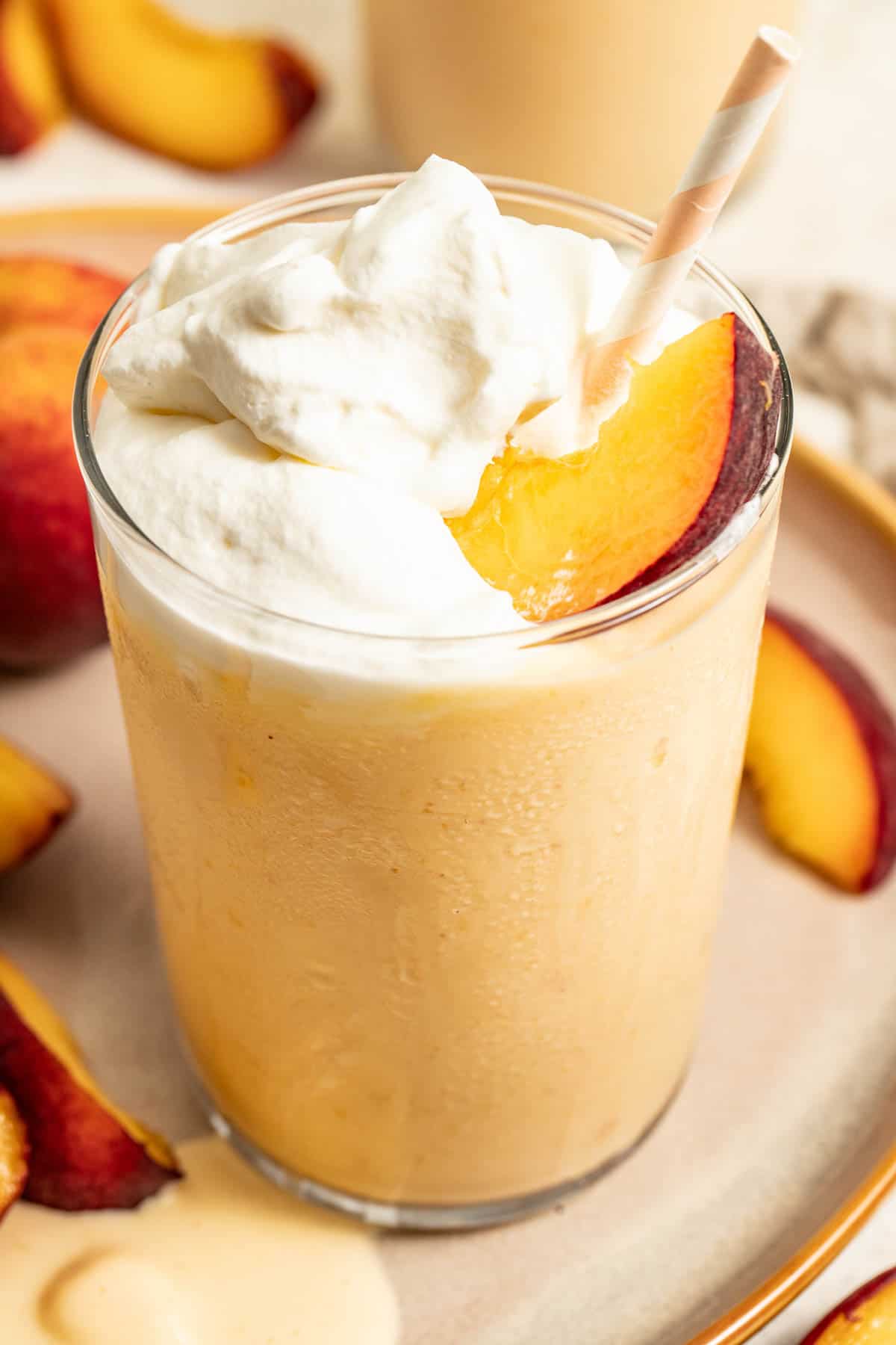 A light yellow milkshake topped with fresh whipped cream, a slice of peach, a straw.