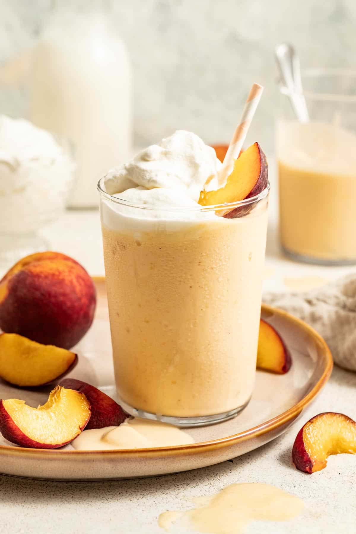 Peach milkshakes topped with fresh peach slices and a striped straw on a table with a glass of milk and vanilla ice cream.