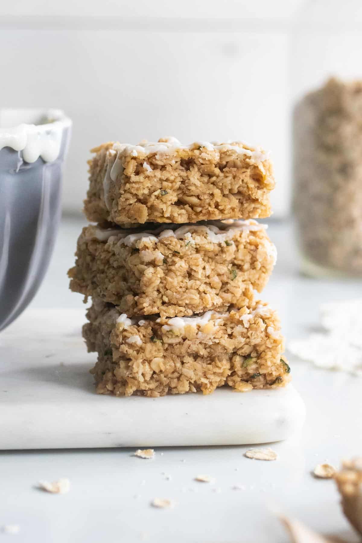 3 zucchini oat bars stacked on top of each other next to a bowl of white cream cheese frosting.