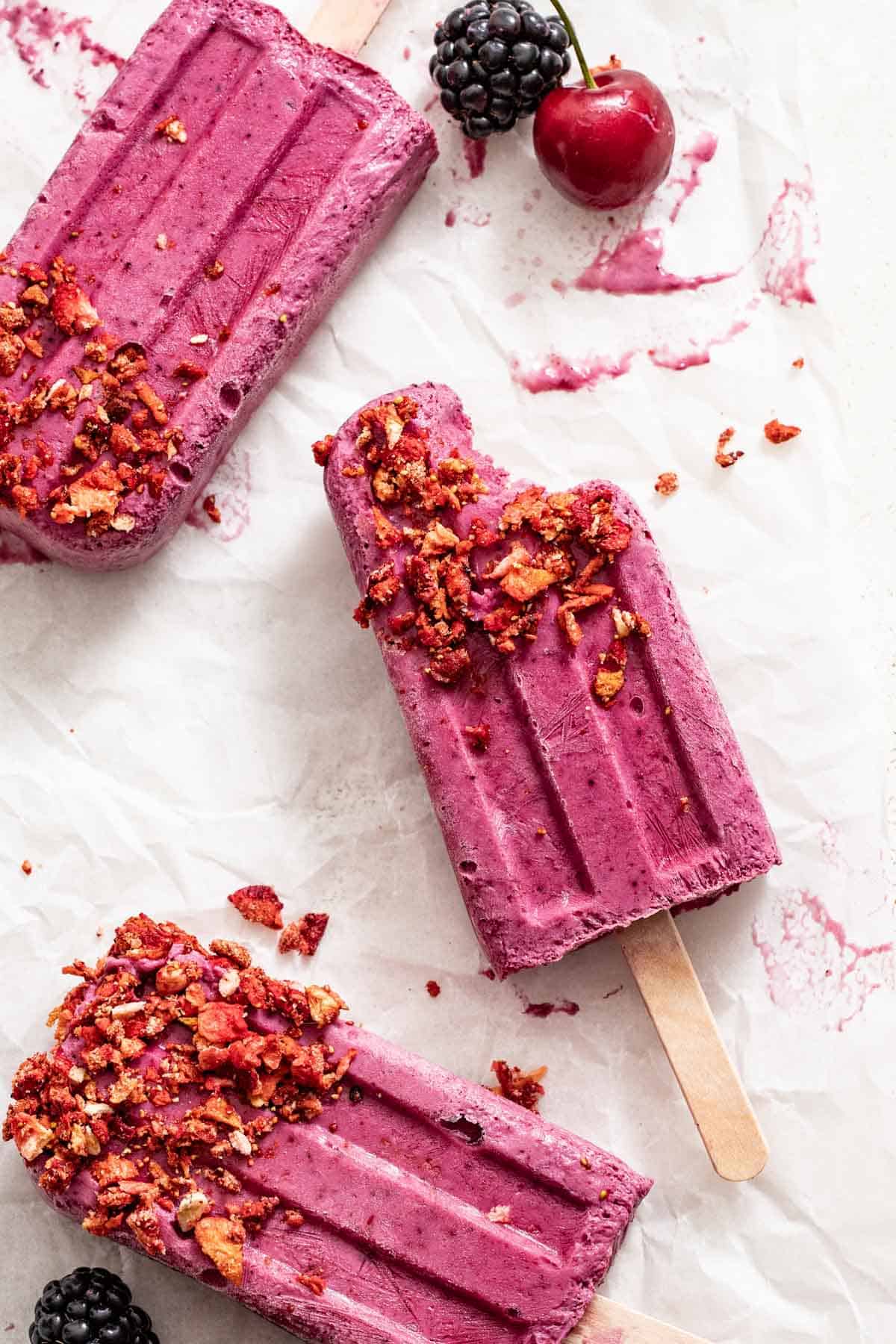 Berry protein popsicles next to cherries and blackberries with a bite out of one of the popsicles.