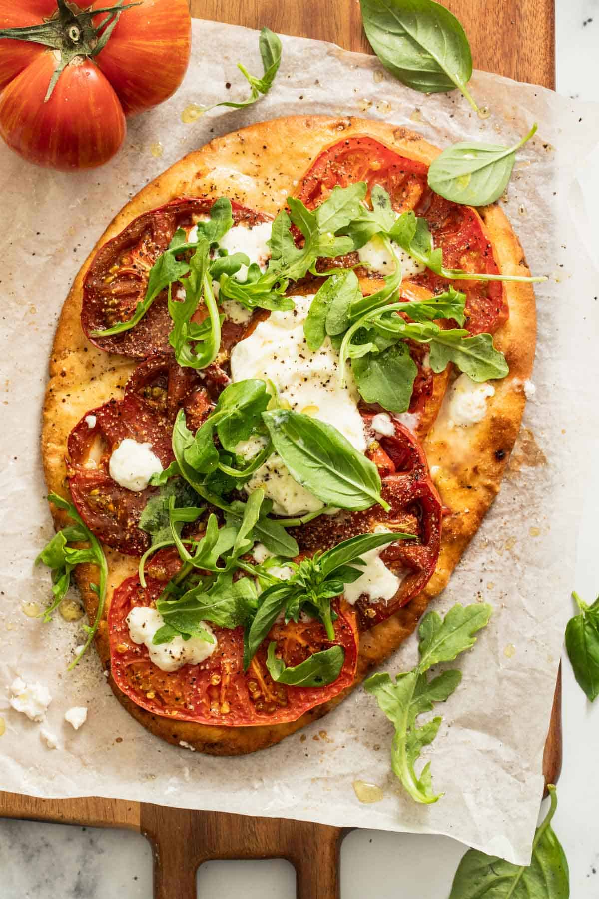 Naan flatbread pizza topped with tomatoes, cheese, and fresh basil on top of a cutting board next to a large heirloom tomato.