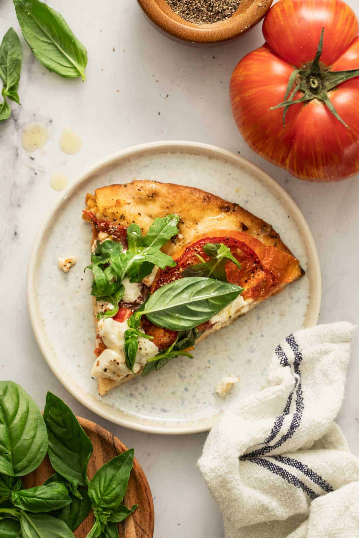 A slice of pizza on a white plate next to tomatoes, a towel, and fresh basil.