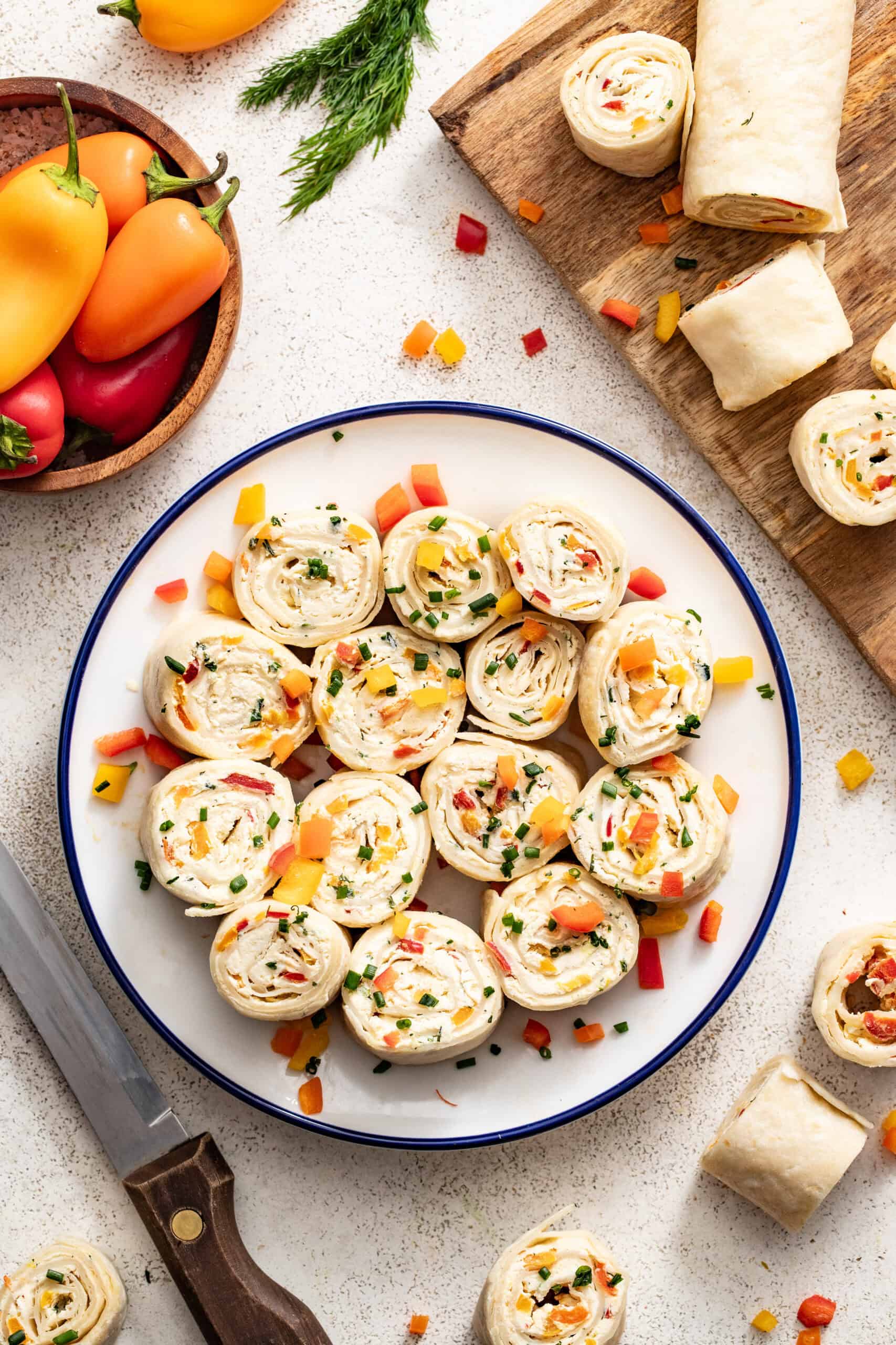 A plate of vegetable tortilla pinwheels with cream cheese and a bowl of peppers next to them.